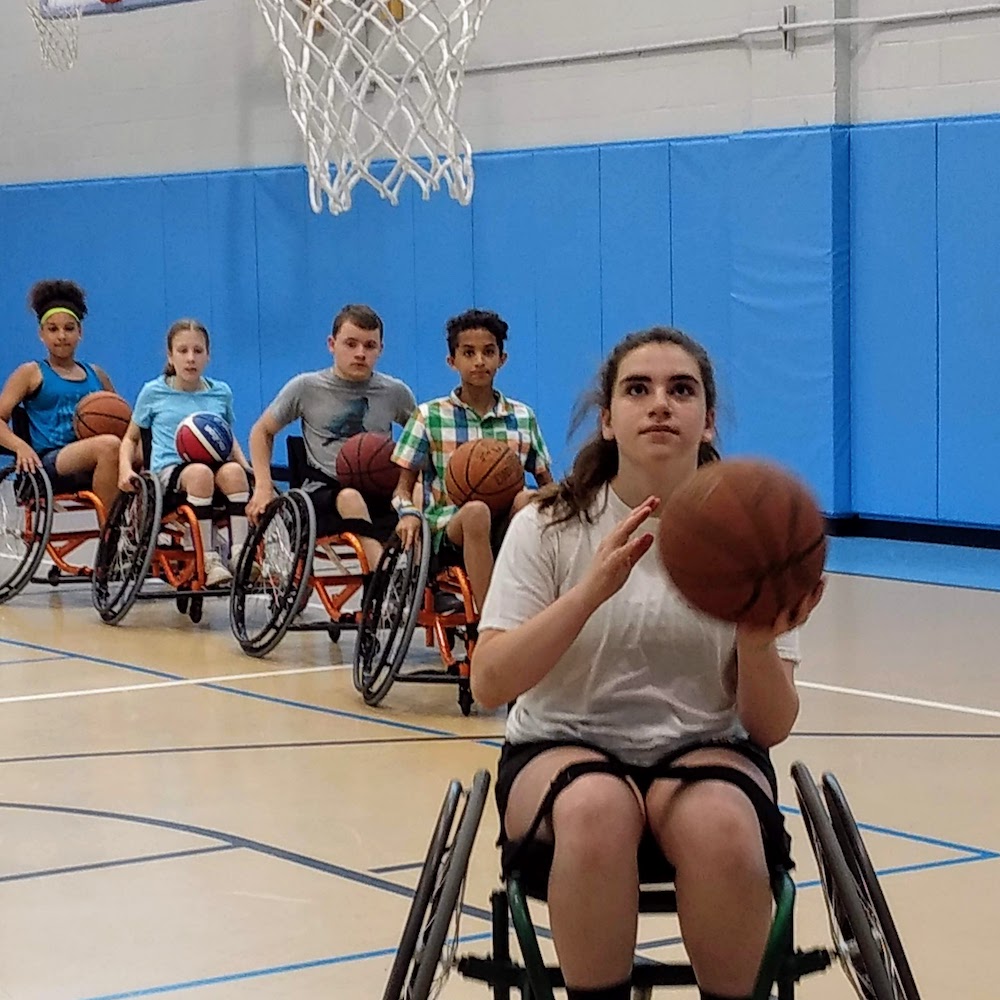 Five youth lined up for layups in a basketball gym, each seated in a sports wheelchair; first youth holds a basketball ready to shoot for a basket.