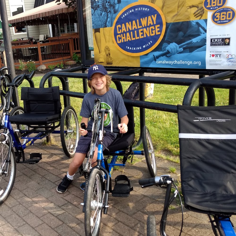 A young boy sits on an adaptive handcycle under a kiosk with a Canalway Challenge banner . Other adaptive cycles are on either side of him.