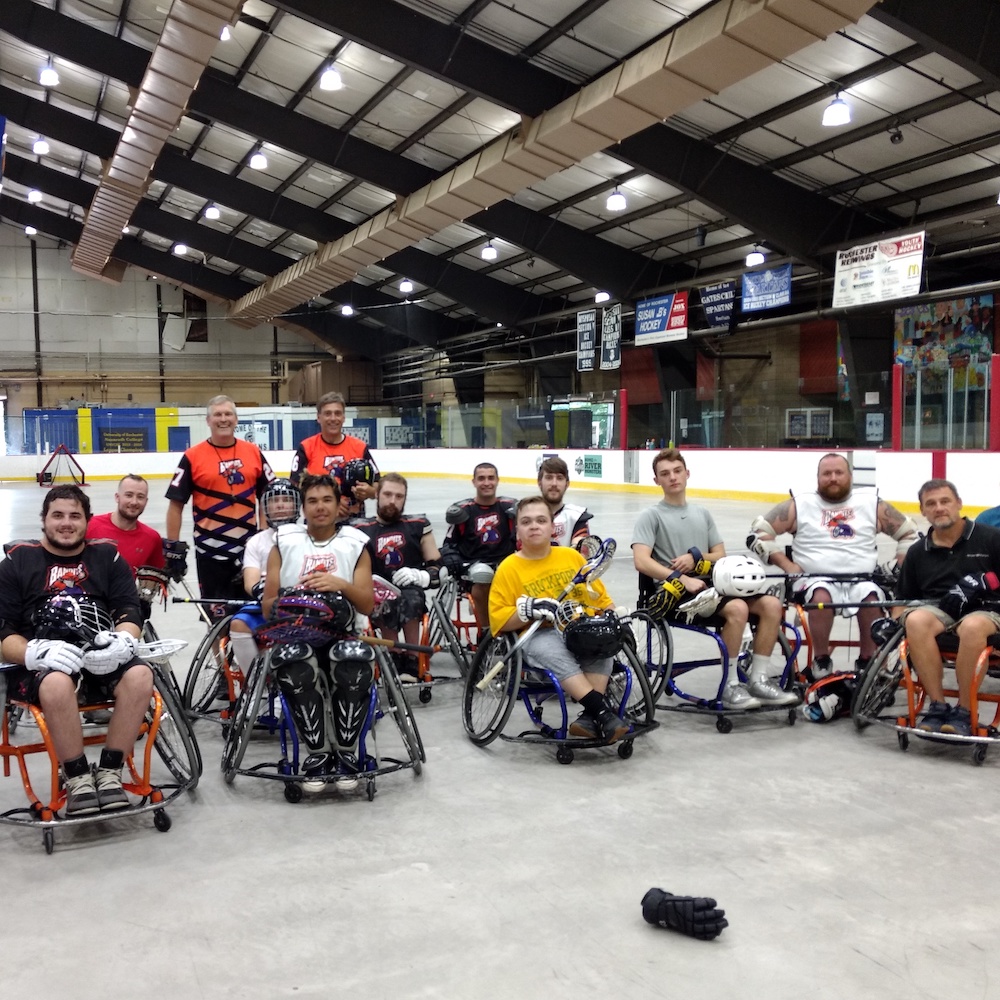 A group of athetes in sports wheelchairs on an indoor lacrosse rink; some players are holding lacross sticks and helmets.