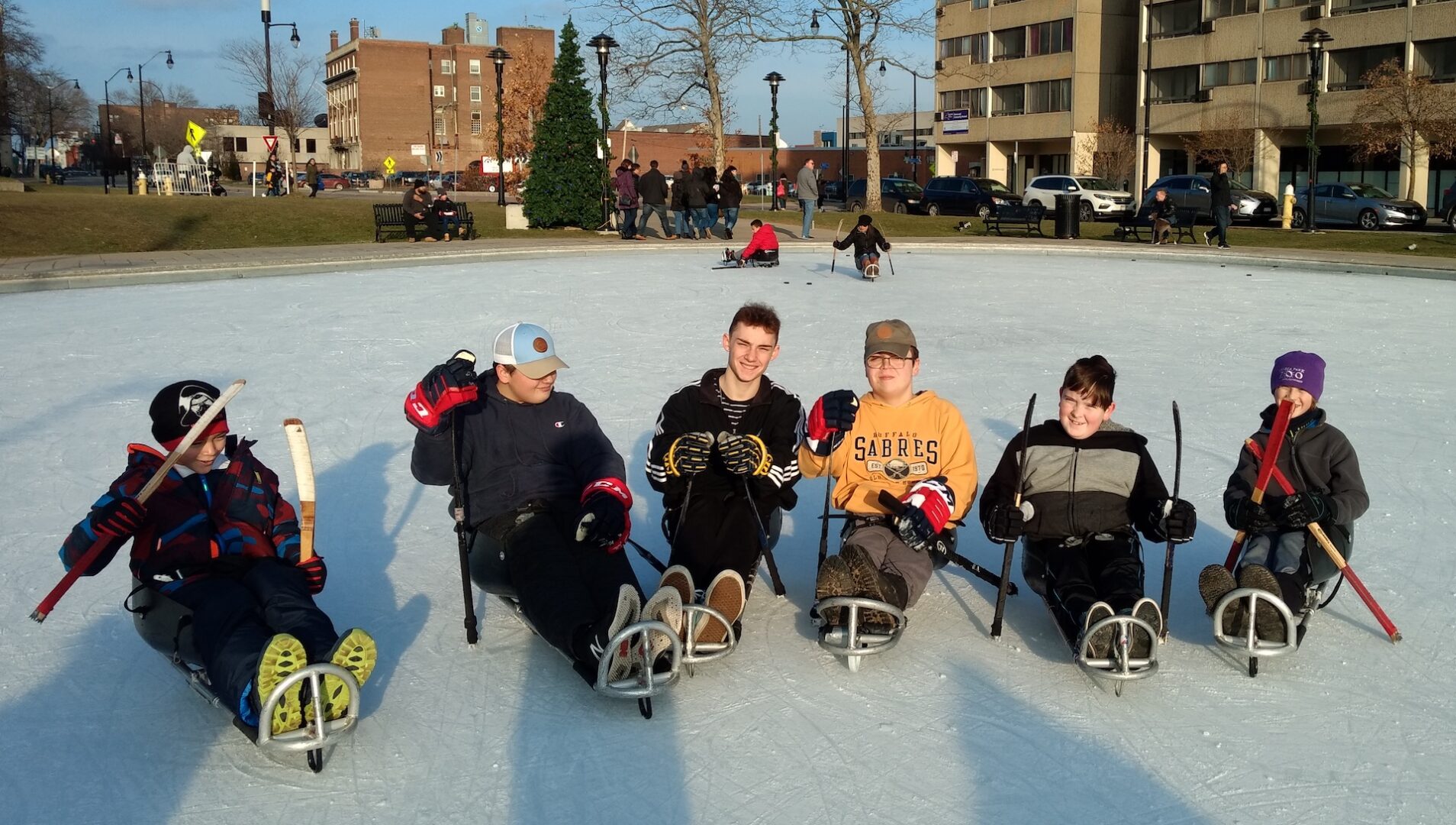 A group of six people in hockey sleds on an outdoor ice rink