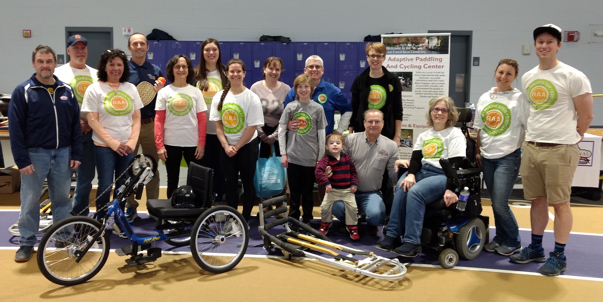 A large group of people is posed for a photo; an adaptive cycle and a hockey sled are in front of the group. Many in the group are wearing RAA tshirts.