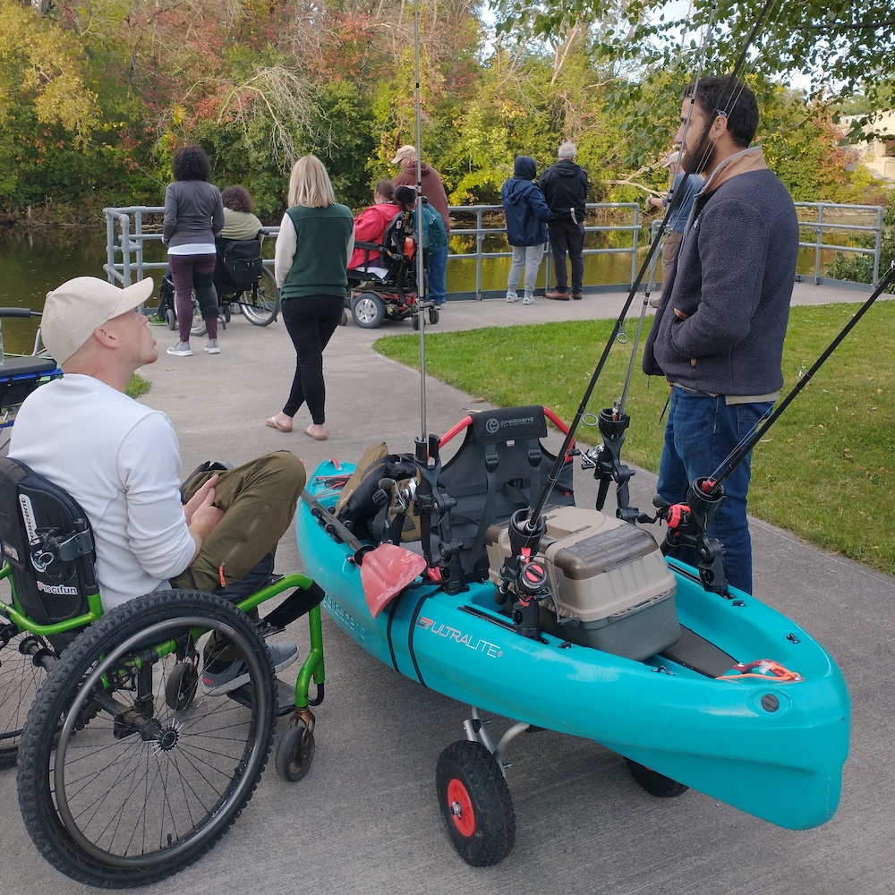 A man sitting in a green manual chair with outdoor tires is talking to a man about the kayak that is outfitted with multiple fishing rods and gear that sits between them. In the background, people are fishing at an accessible fishing railing by a river.