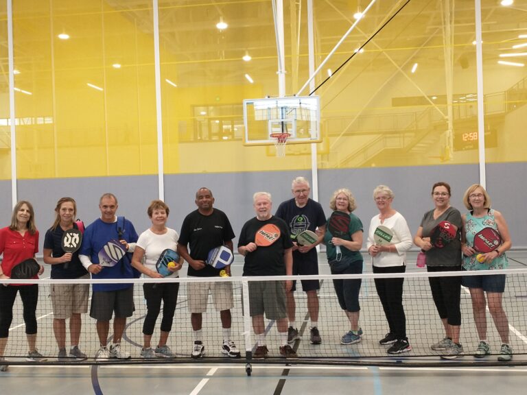 A group of 11 people lined up behind a pickleball net in an indoor gym. Each is holding a pickleball paddle and smiling.
