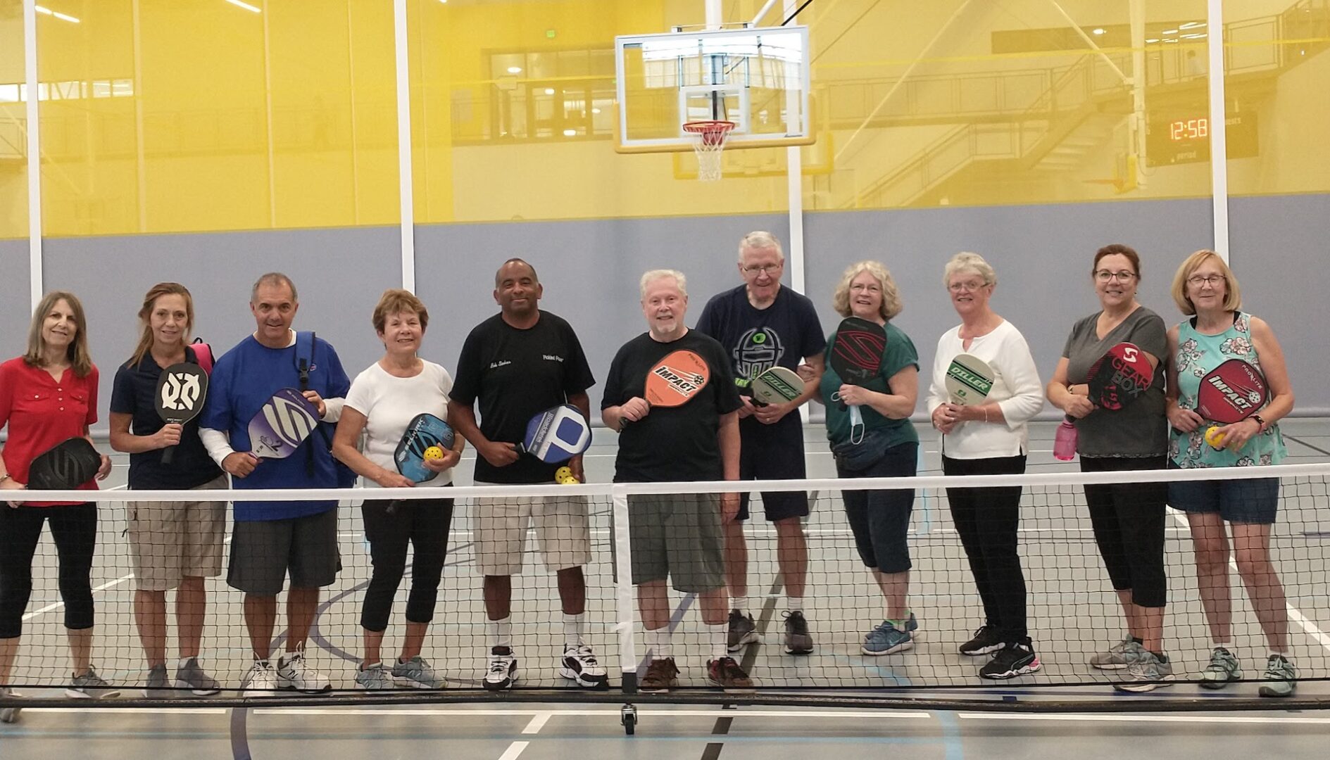 A group of 11 people are lined up behind a pickleball net in an indoor court.. Each is holding a pickleball paddle and/or pickleball, and smiling