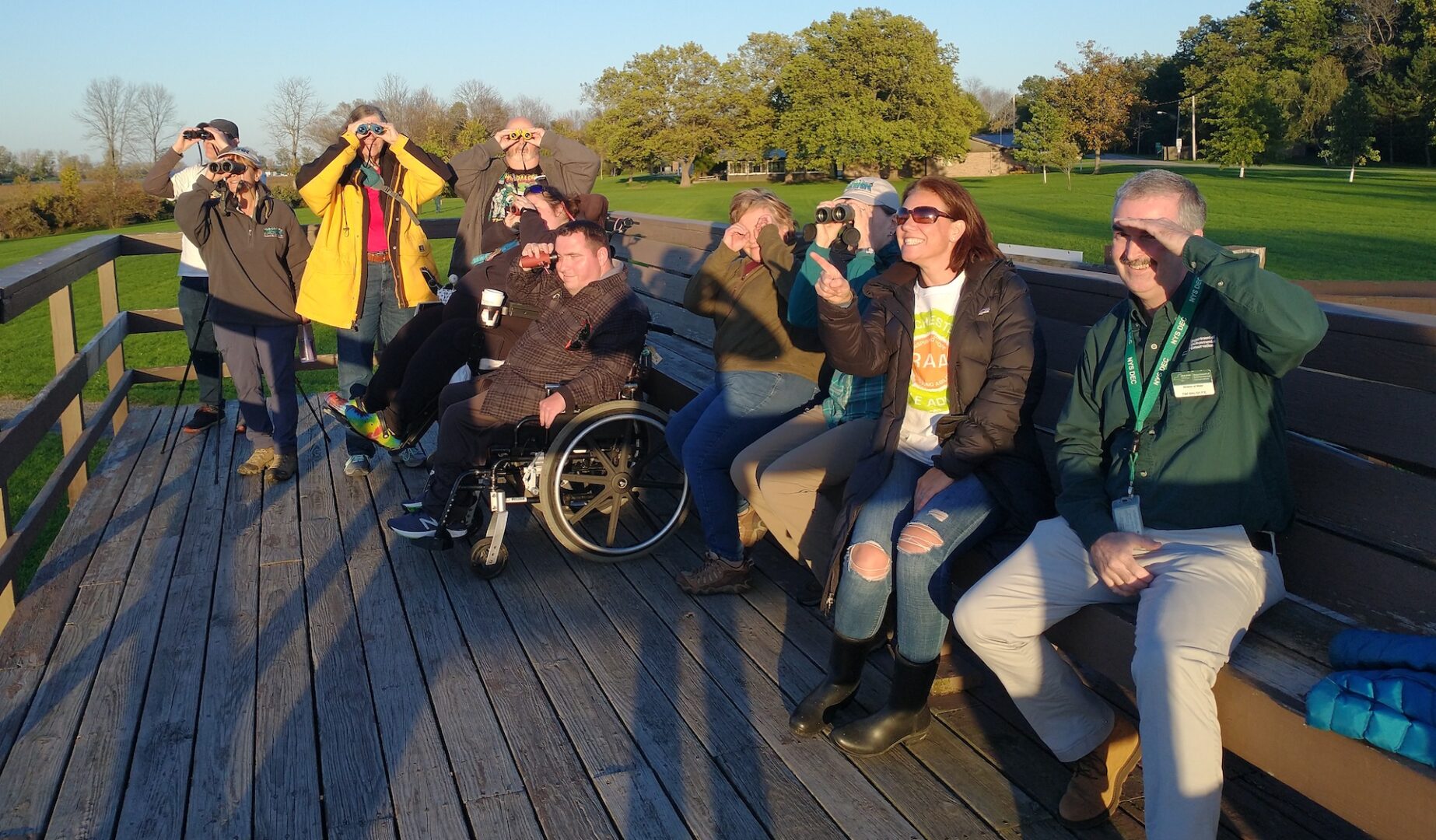 A group of 10 people stands or sits on a raised nature observation deck with grassy expanse around them and trees behind them. Two are in wheelchairs. All are holding binoculars, monoculars or just looking bared-eyed.