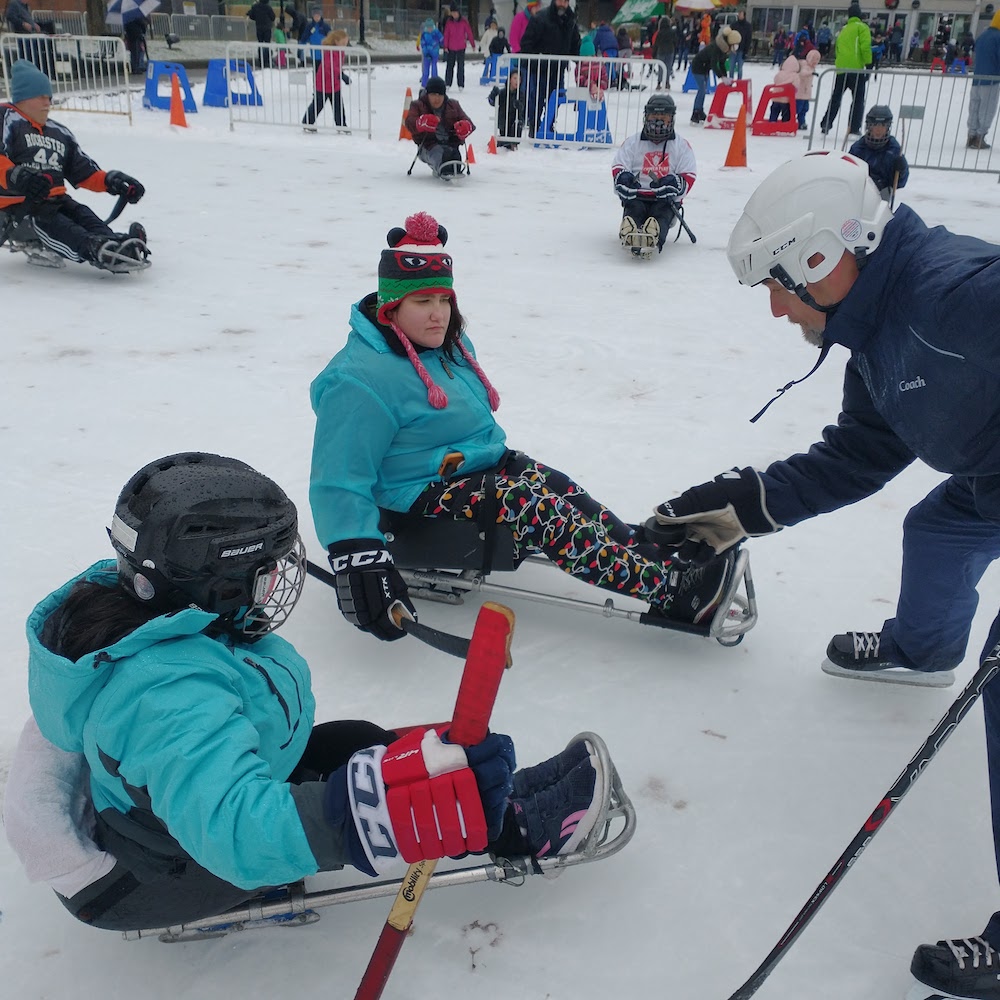 An outdoor ice rink with skaters in the background. Two sled hockey players are on sleds with sticks, gloves and helmets waiting for a man to drop the puck to start the game.