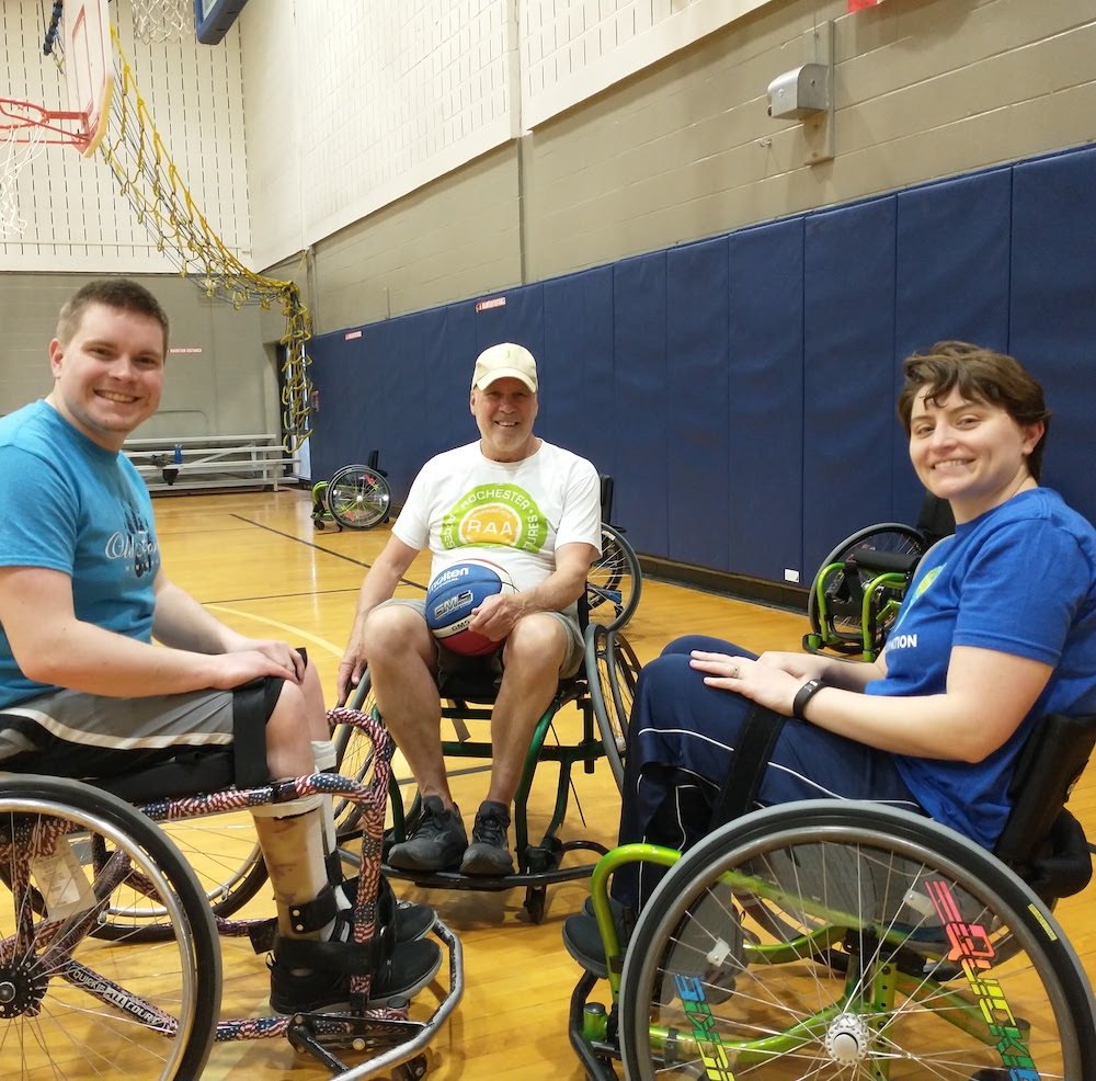 Three adults are sitting in sports wheelchairs in a gym; one holds a basketball on his lap. One is wearing an RAA tshirt.