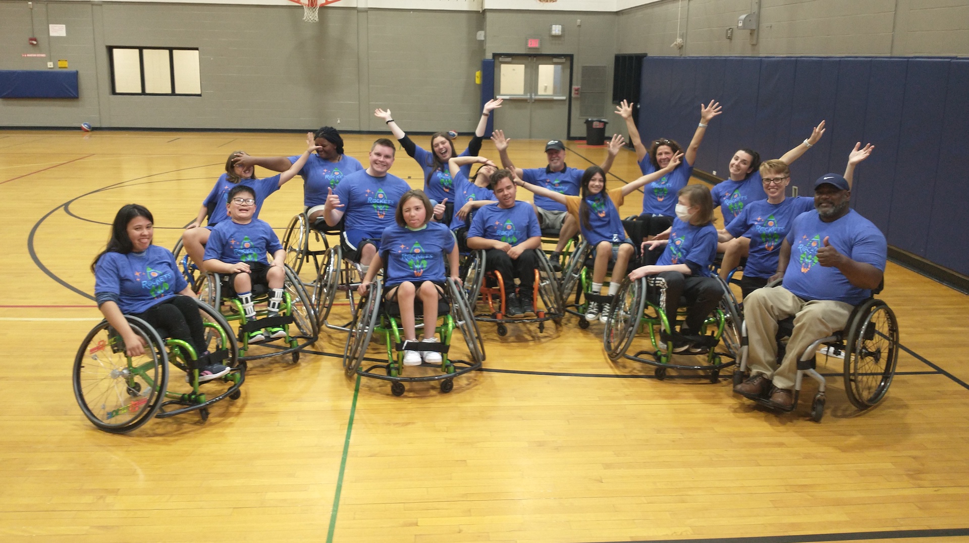 A group of youth and adults in a basketball gym, all in sports wheelchairs and blue tshirts with a rocket graphic on them; all in various poses for a "funny photo shot.