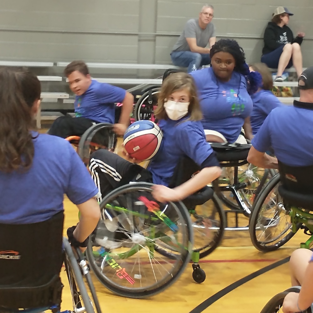 Action shot of youth in sports wheelchairs playing basketball; center youth holds the basketball in one hand, the other hand is on the wheel as he looks back over his shoulder.