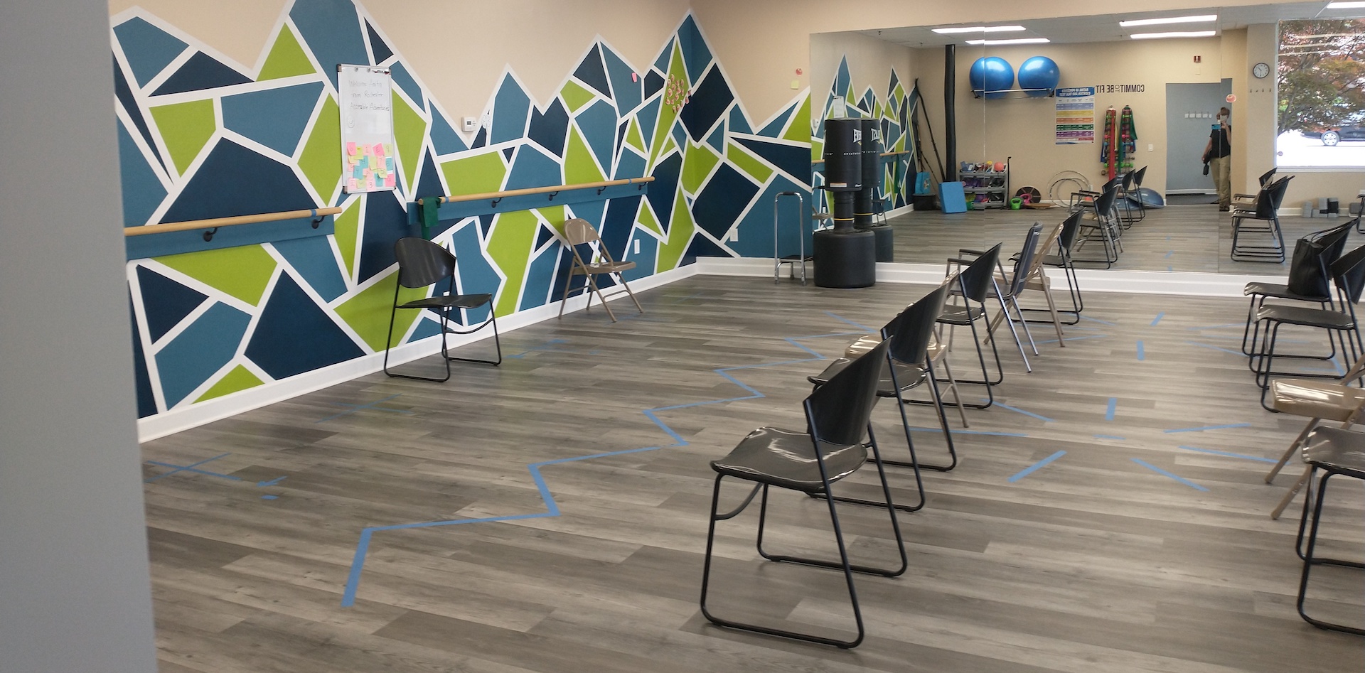 A spacious therapy gym has an artistic cubicle artwork on the wall, two rows of chairs face that wall; a floor-to-ceiling mirro extends along the side wall, showing therapy balls and equipment on the other wall. Taped paths on the floor indicated placement for various physical therapy exercises.