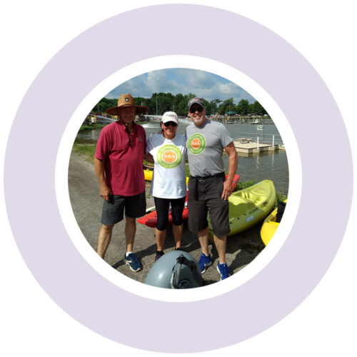 A light purple graphic ring encircles a photo of three people standing in front of kayaks; a wide water area on the Erie canal is behind them. Two of them are wearing RAA tshirts.