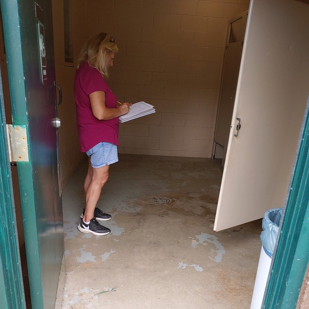 A person is standing in a park bathroom, doors propped open. She holds a clipboard and is writing on it.