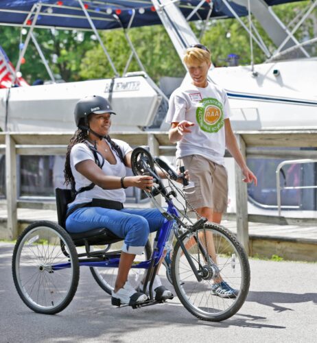 A woman is pedaling a handcycle on a paved path; a woman in an RAA shirt is walking beside her pointing at the steering mechanism. 