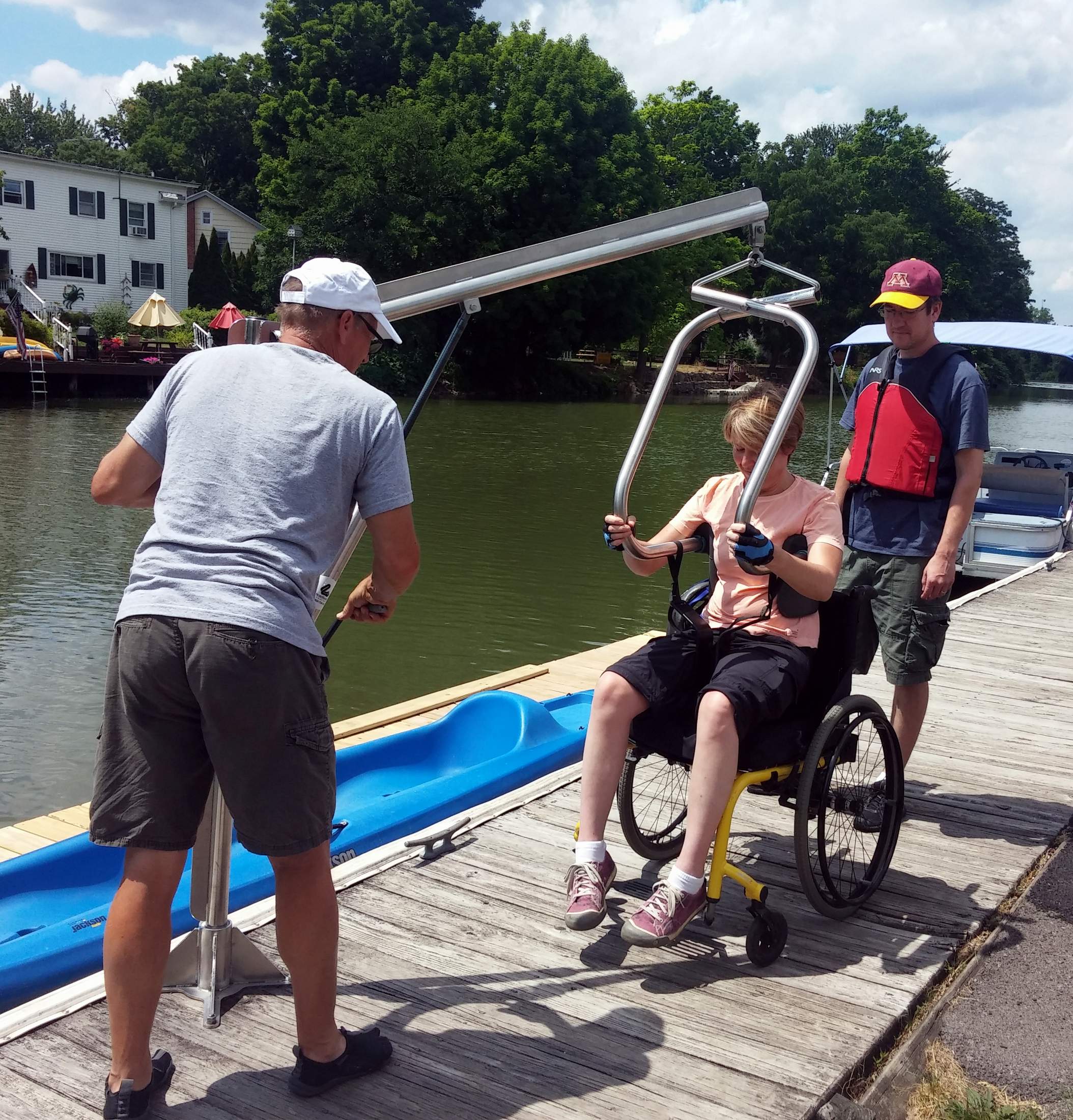 A woman sits in her manual wheelchair and has the arms of a hoyer lift (mounted on a dock) under her shoulders. A man is pumping the hydraulic lift to transfer her into a blue kayak that is waiting on a side dock. A man stands behind the woman in a lift jacket.