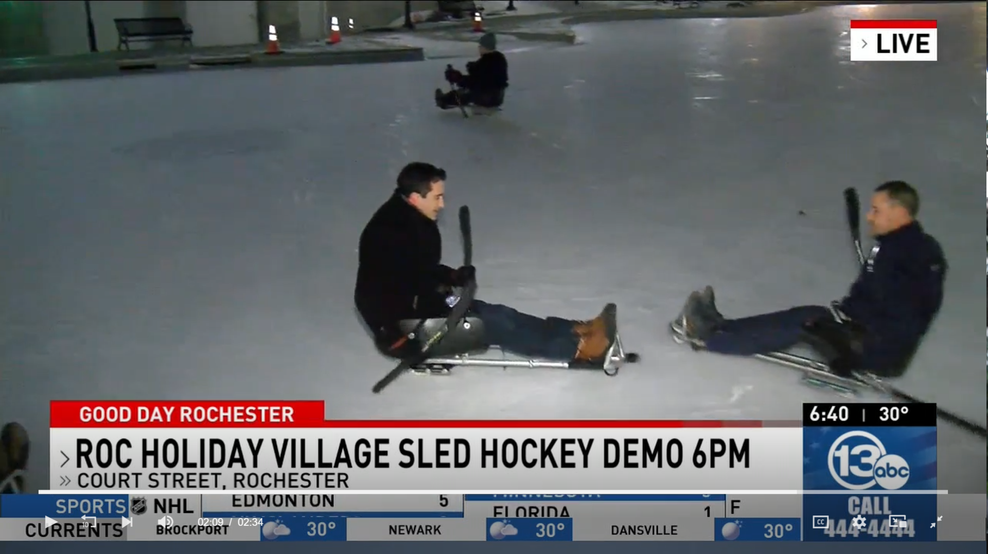 A screenshot of a TV news screen with "ROC Holiday Village Sled Hockey Demo"; two people on hockey sleds with sticks are moving towards a puck on an outdoor ice rink