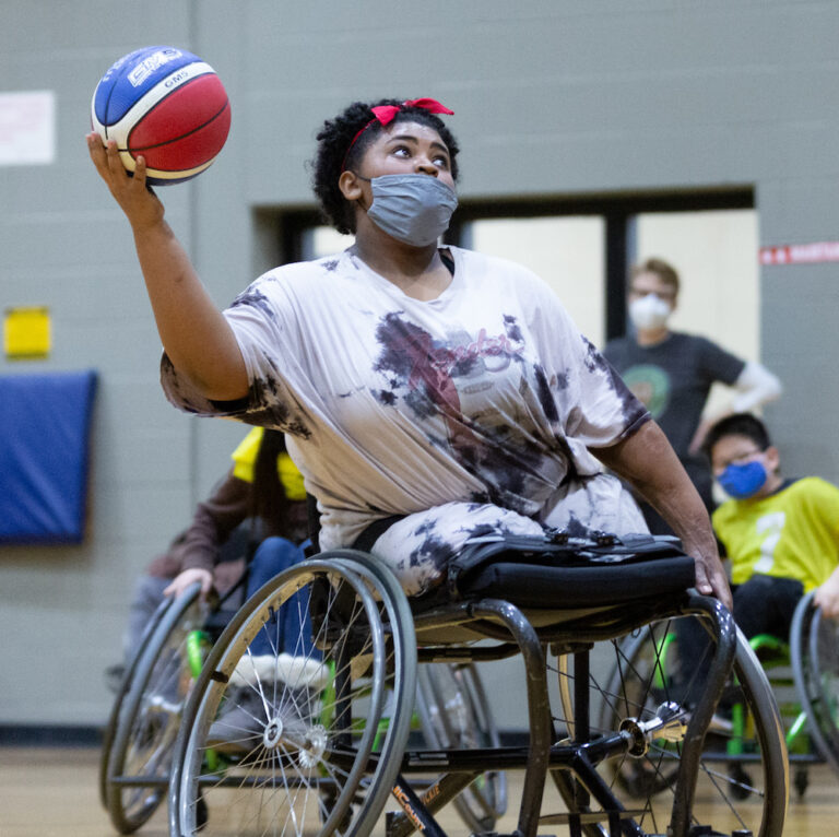 A closeup of a youth with bi-lateral amputation in a black sports chair, one hand holding a basketball, eyes focused on making a shot;