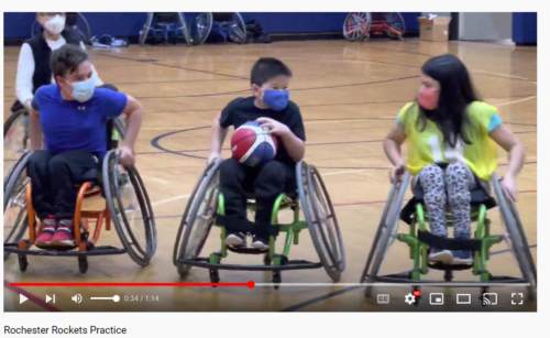 Three youth in sports chairs push down a gym basketball court, one with ball in hand.