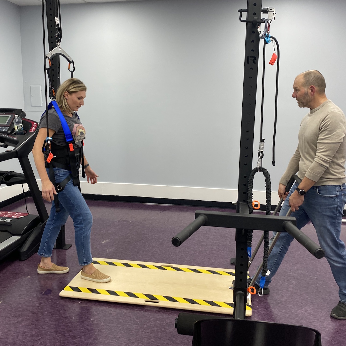 A woman has on a harness that is suspended from a metal bar above her. One foot is on a board on the floor. A man holds on to a handle that is attached to the Slip Board. He is pulling the board, which will cause the woman in the harness to experience slipping - and helping her body learn to respond safely to the recovery.