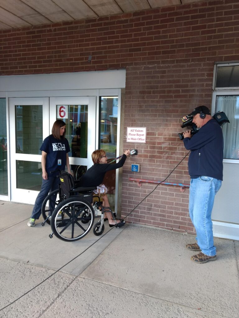A woman is seated in a manual wheelchair and is reaching her keytag on a lanyard around her neck towards the door opener sensor; she is just out of reach of the sensor. A man with a video camera is recording.