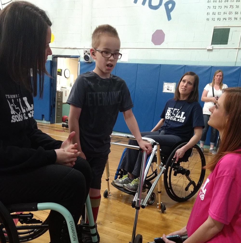 Boy with walker playing in a school gym; He is talking to a woman who is on her knees on the gym floor, and to another woman in a manual wheelchair. A third woman is seated in a manual wheelchair listening.