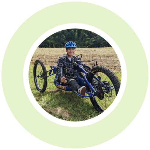 A graphic light green ring encircles a youth sitting on an adaptive hand-cranked mountain bike on the grass