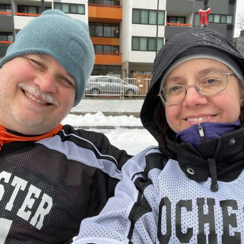 Close up of a man and woman in hockey helmets and jerseys, on an outdoor ice rink, smiling