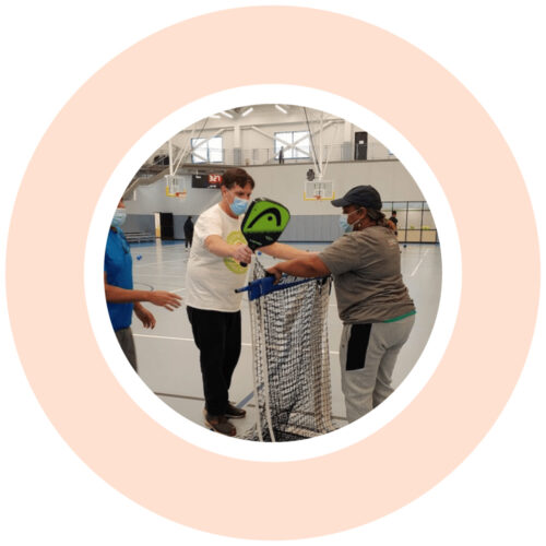 A light orange graphic circle around an image of three people setting a pickleball net and paddles.