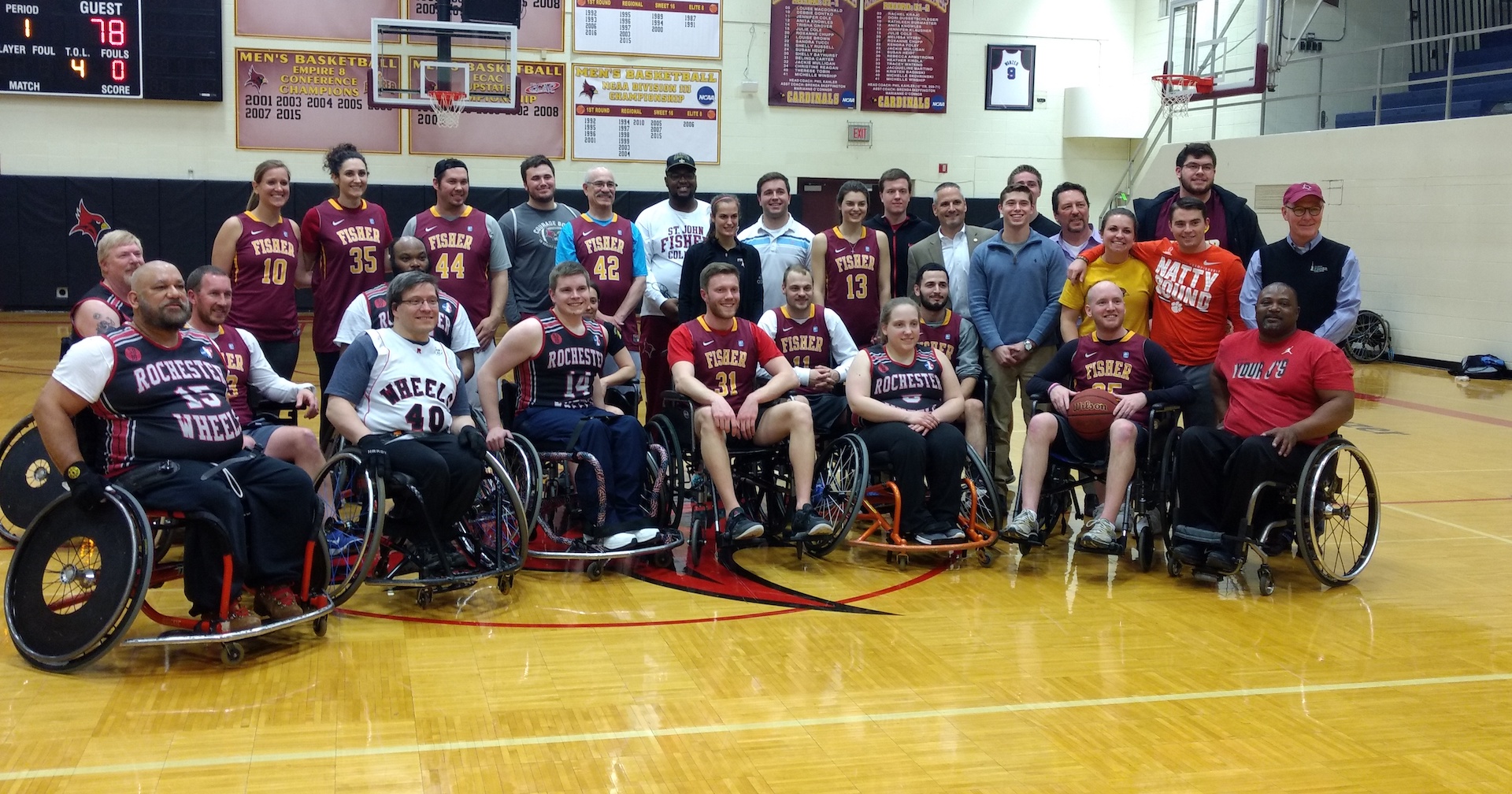 A large group of students with Fisher jerseys stands behind a group of athletes in sports chairs wearing "Rochester Wheels" jerseys; gym walls posted with school sports information