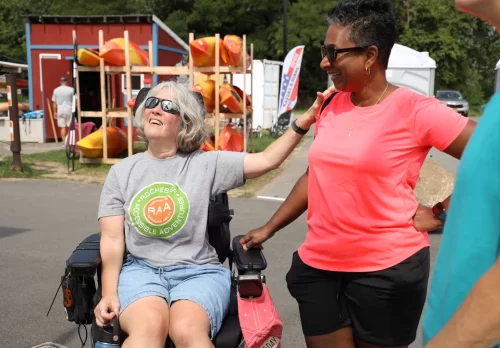 Two women smiling broadly in front of a rack of kayaks. One woman with short white and black hair in tshirt and shorts is seated in a power chair. Her hand rests on the other person's shoulder as that person stands beside her in bright orange tshirt and black shorts, hand resting on her power chair.
