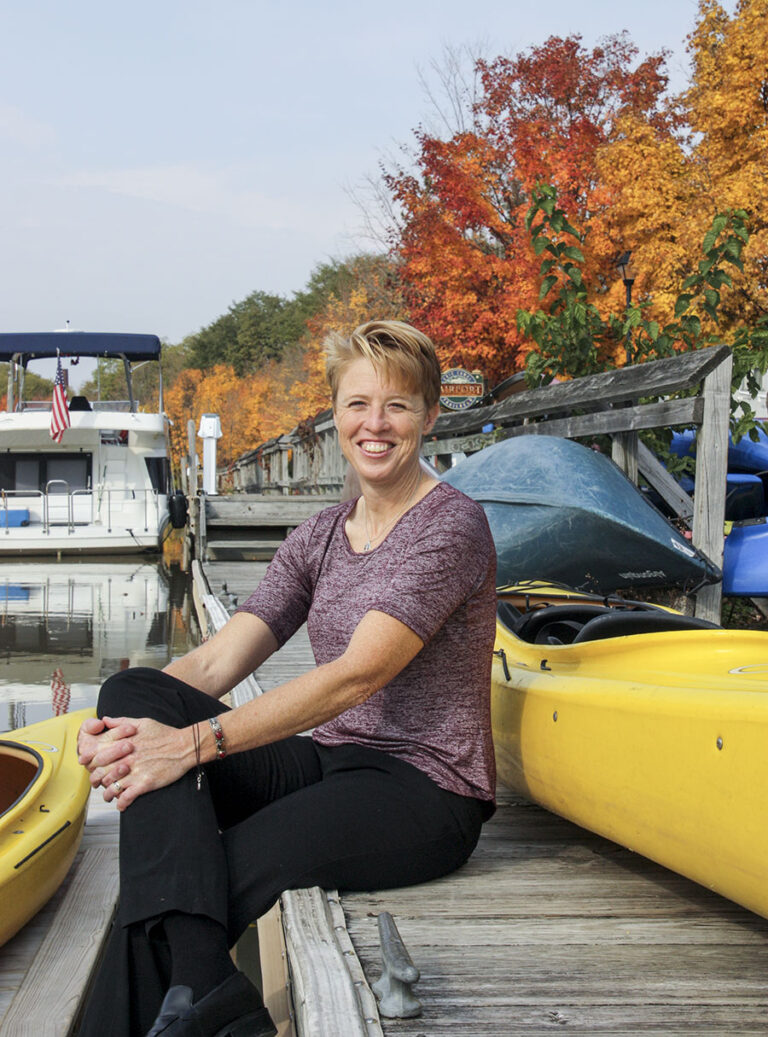 A woman with short red hair sits on a dock along the Erie Canal; yellow and blue kayaks are behind her; a houseboat is docked in the background.