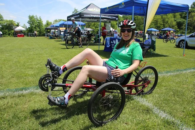 A woman in a green tshirt and shorts with a bike helmet on smiles as she demonstrates an adaptive three-wheeled cycle. Vendor tents are set up in the background on a grassy area.