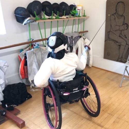A person seated in a wheelchair, dressed in fencing shirt and helmet, posed with a weapon at an artistic rendering of a fencer on the wall