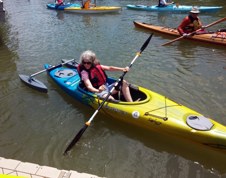 A woman paddles a yellow and blue kayak. The kayak has short outriggers at the back; there is a paddle pedestal that support the paddle.
