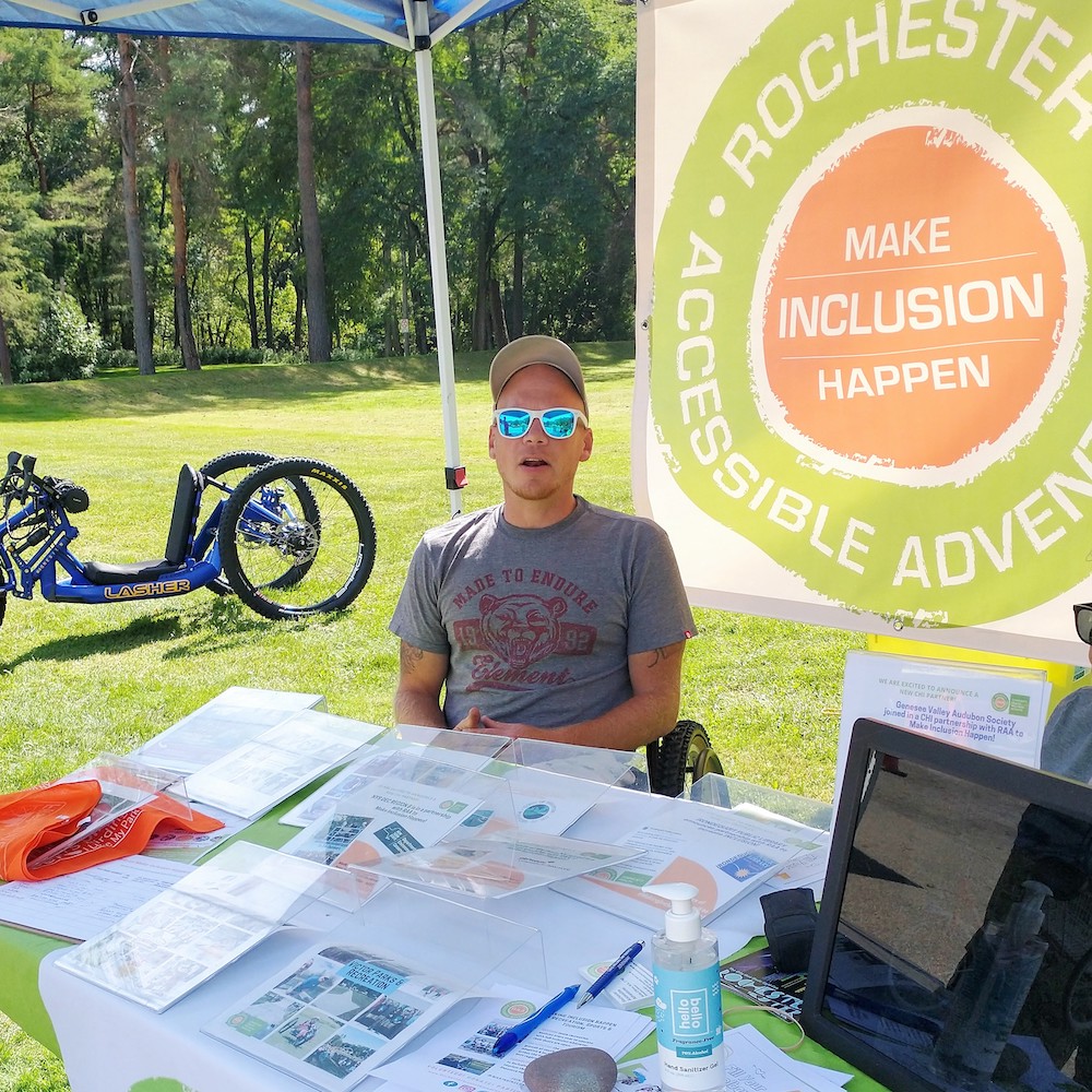A man with sunglasses and a ballcap sits behind a table full of information flyers; a large white, green and orange banner hangs behind him "Rochester Accessible Adventures: Make Inclusion Happen". An adaptive mtn bike is in the background on the grass. The man is seated in a manual wheelchair.