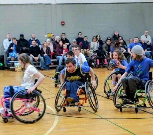Youth in sports chairs race up a gym court in a wheelchair basketball game; fans cheer in bleachers behind them