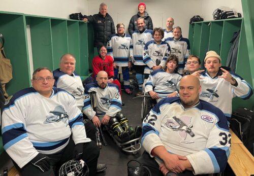 A group of people in mostly white and blue hockey jerseys pose for a picture in a locker room, in wheelchairs, on hockey sleds, or standing.