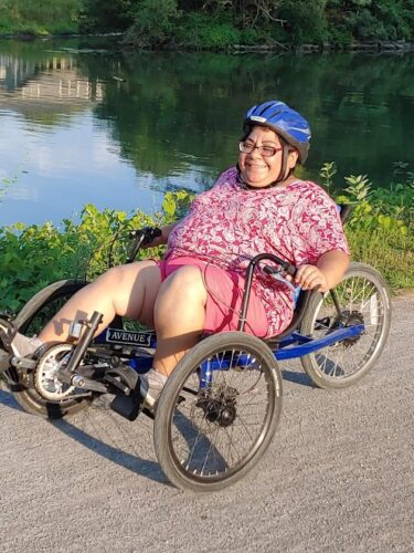 A woman wearing pink shorts and a pink print shirt with a blue bike helmet rides a three-wheeled cycle on a crushed stone pathway alongside the Erie Canal.
