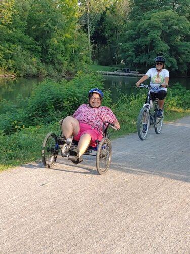 A woman wearing pink shorts and a pink print shirt with a blue bike helmet rides a three-wheeled cycle on a crushed stone pathway alongside the Erie Canal. A woman in a white RAA shirt rides a two wheeled bike along with her.
