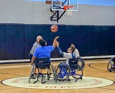 Two youth in sports chairs are set for the tip off of the wheelchair basketball game. A referee has tossed the ball in the air above them and they both are reaching for the ball.