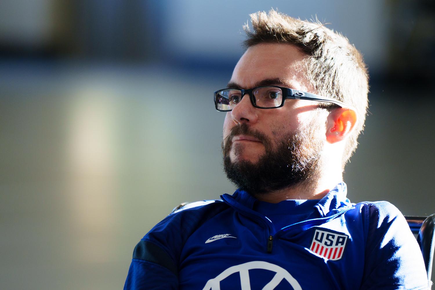 Headshot of a man with short dark hair and beard, with dark rimmed glasses and wearing a blue USA Team shirt