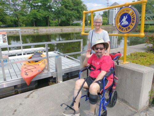 A couple smiles by the adaptive kayak launch at Brockport Welcome Center. He is seated in a wheelchair and she is standing beside him, with an orange kayak on the dock behind them.