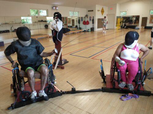 Two people seated in chairs get set to spar in a fencing studio, each wearing a fencing helmet