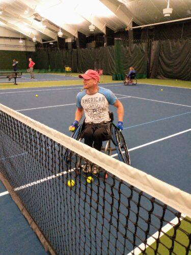 A person is seated in a sports chair behind a tennis net ready to volley over the net