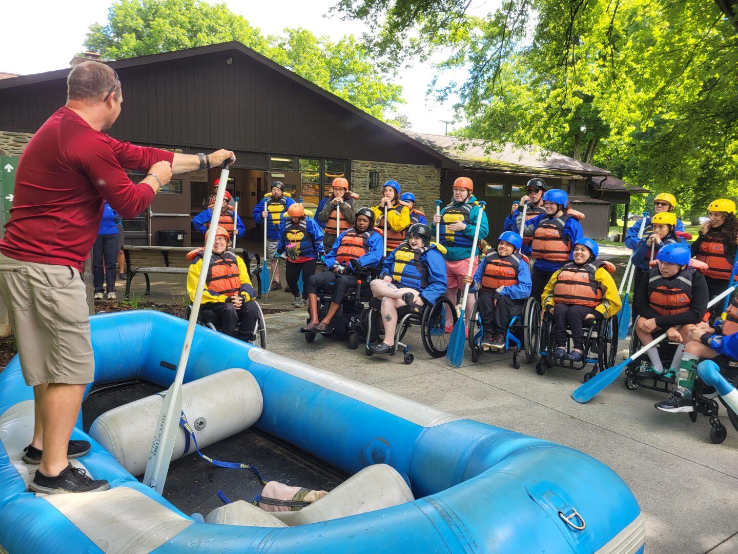 A rafting leader is standing on a whitewater raft on dry ground, explaining details to rafting to the group of people in the background, a mix of people seated in wheelchairs and standing.