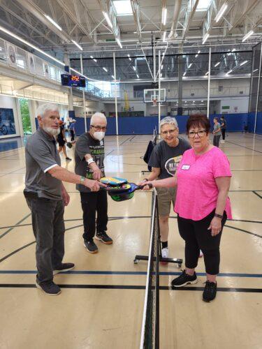 Four people stand two each on a pickleball court in a gym, reaching across the net to join paddles at the end of a game.