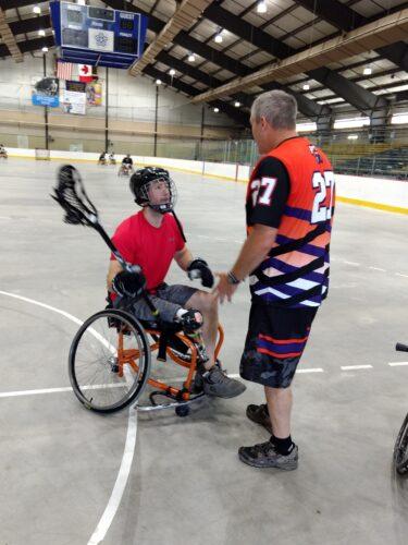 A person sits in a sports chair in lacrosse gear talking with a coach standing beside him.