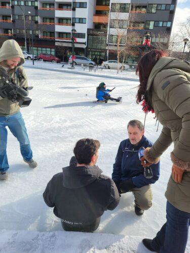 A tv crew is on an outdoor skating rink. The camara person is standing out on the ice filming as a coach helps a tv reporter into a hockey sled; another reporter is holding a microphone to pick up what the coach is saying.