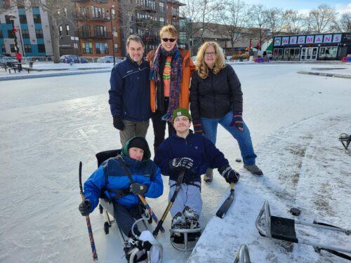 Several people on an outdoor ice rink, two are in hockey sleds and three are standing behind them