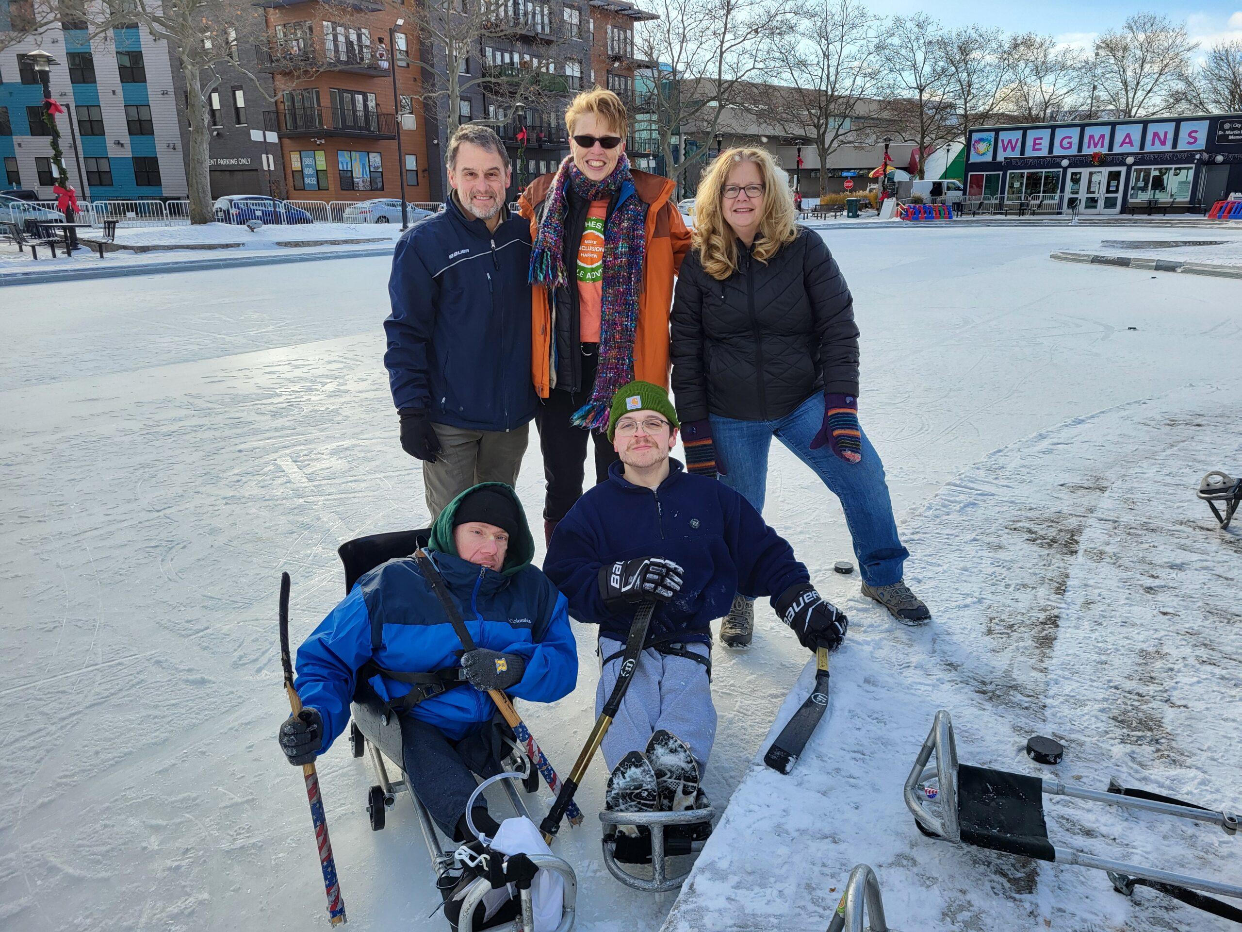 Five people pose for a photo on an outdoor skating rink at Roc Holiday Village. Two sled hockey players on sleds, with sticks in hand. Three people stand behind them.