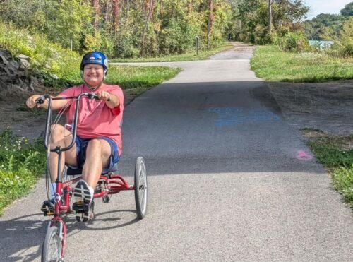 A man on a three-wheeled red bike with upright seat and handlebars. He is smiling and is on an Erie Canalway path.